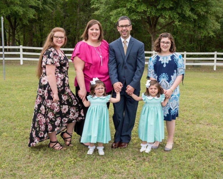 Family of seven smiling outdoors. Two young girls in matching dresses hold hands with parents.