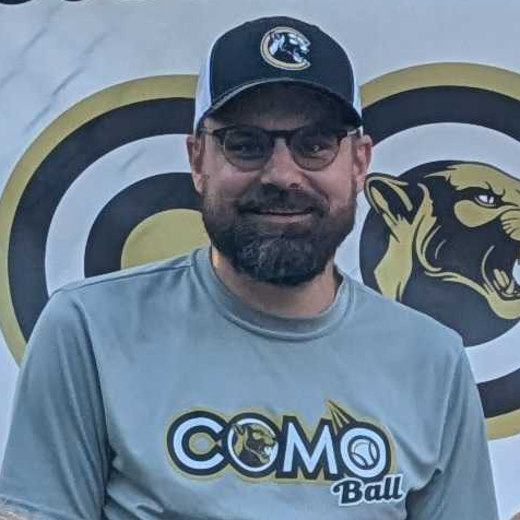 Man in green polo stands on a football field, arms crossed, stadium in background.