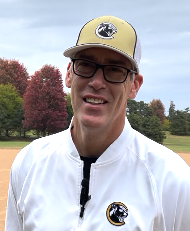 Football coach with arms crossed, standing on a football field. He wears a navy polo shirt and gray pants.