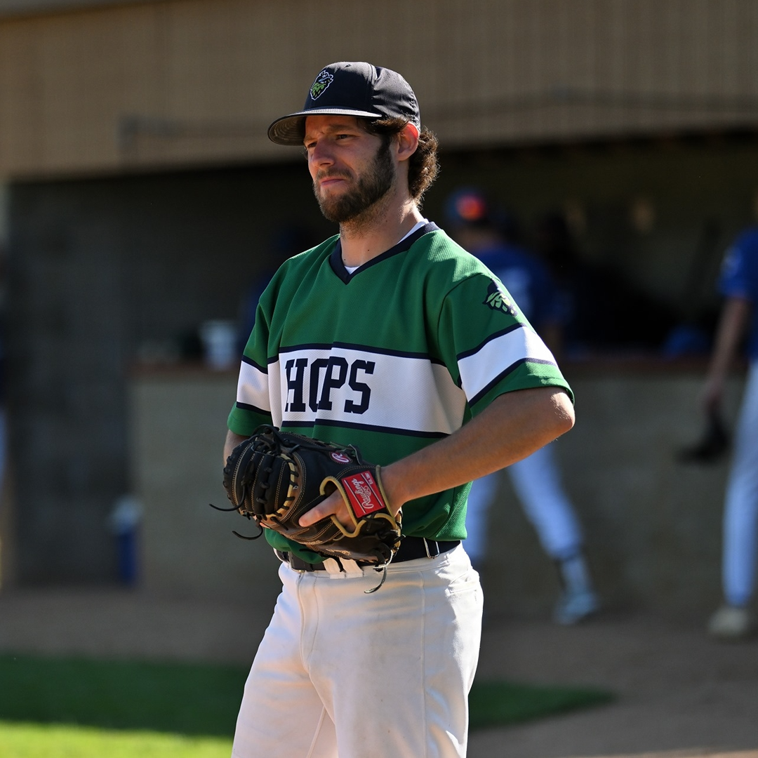 Football coach with arms crossed, looking stern on a sunny field, players in the background.