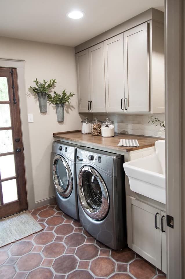 A laundry room with gray cabinets, a wooden counter, silver appliances, a white farmhouse sink, and brown hexagon tiles.
