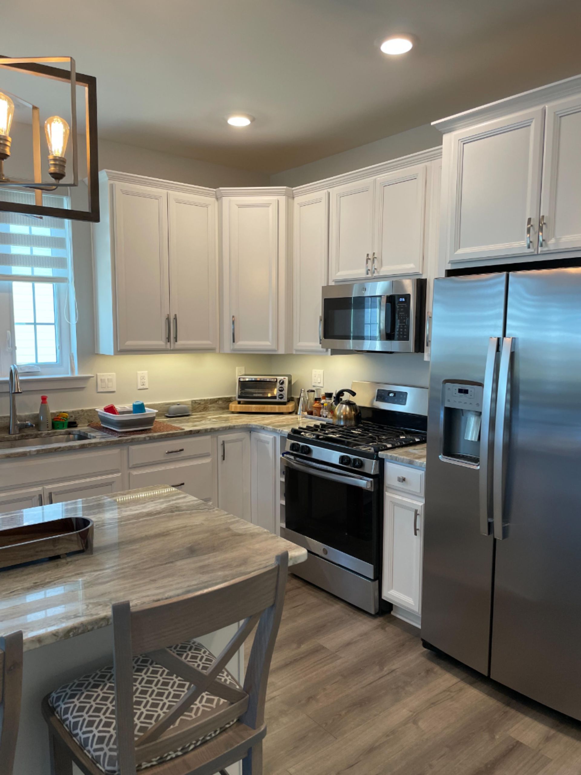A modern kitchen with white cabinets, stainless steel appliances, a light-colored granite countertop, and a wooden chair.