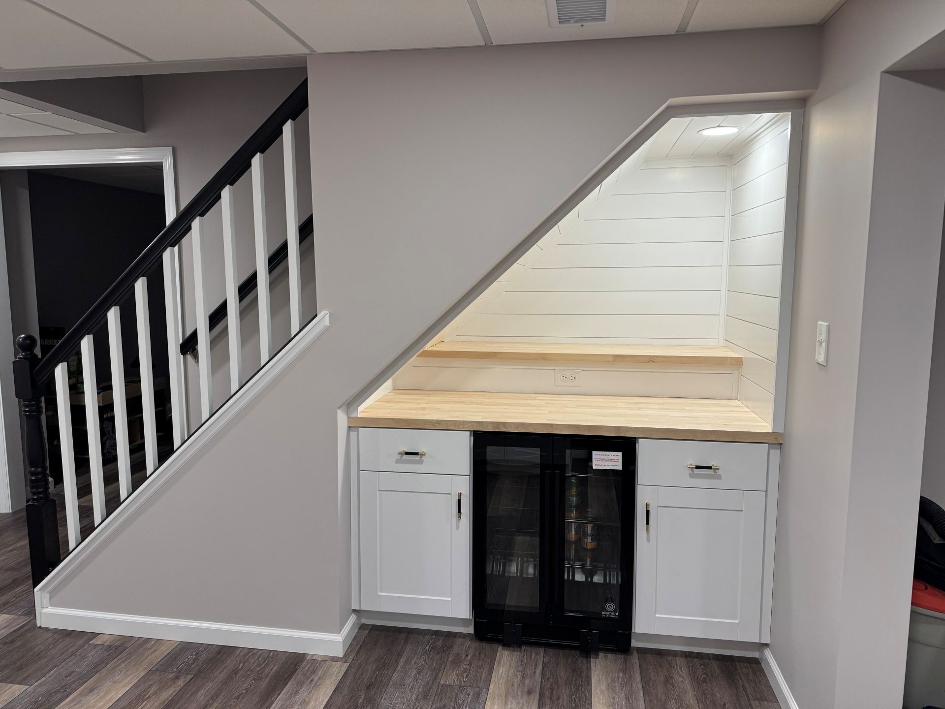 A custom basement bar built under a staircase, featuring white cabinets, a glass-door beverage cooler, and wood shelving.