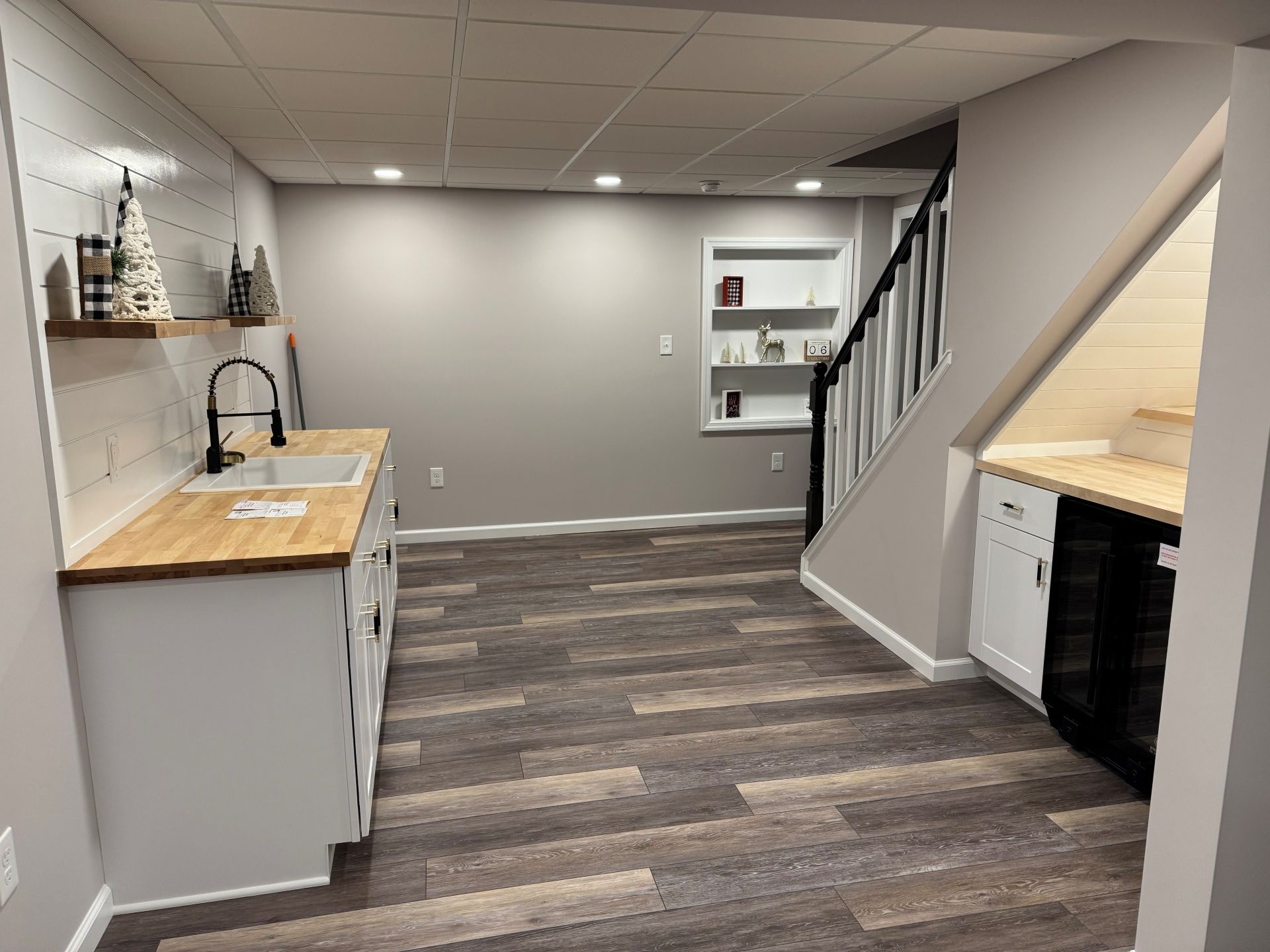 A basement kitchen area with grey cabinets, wood countertops, white shiplap walls, recessed shelving, and wood-look floors.