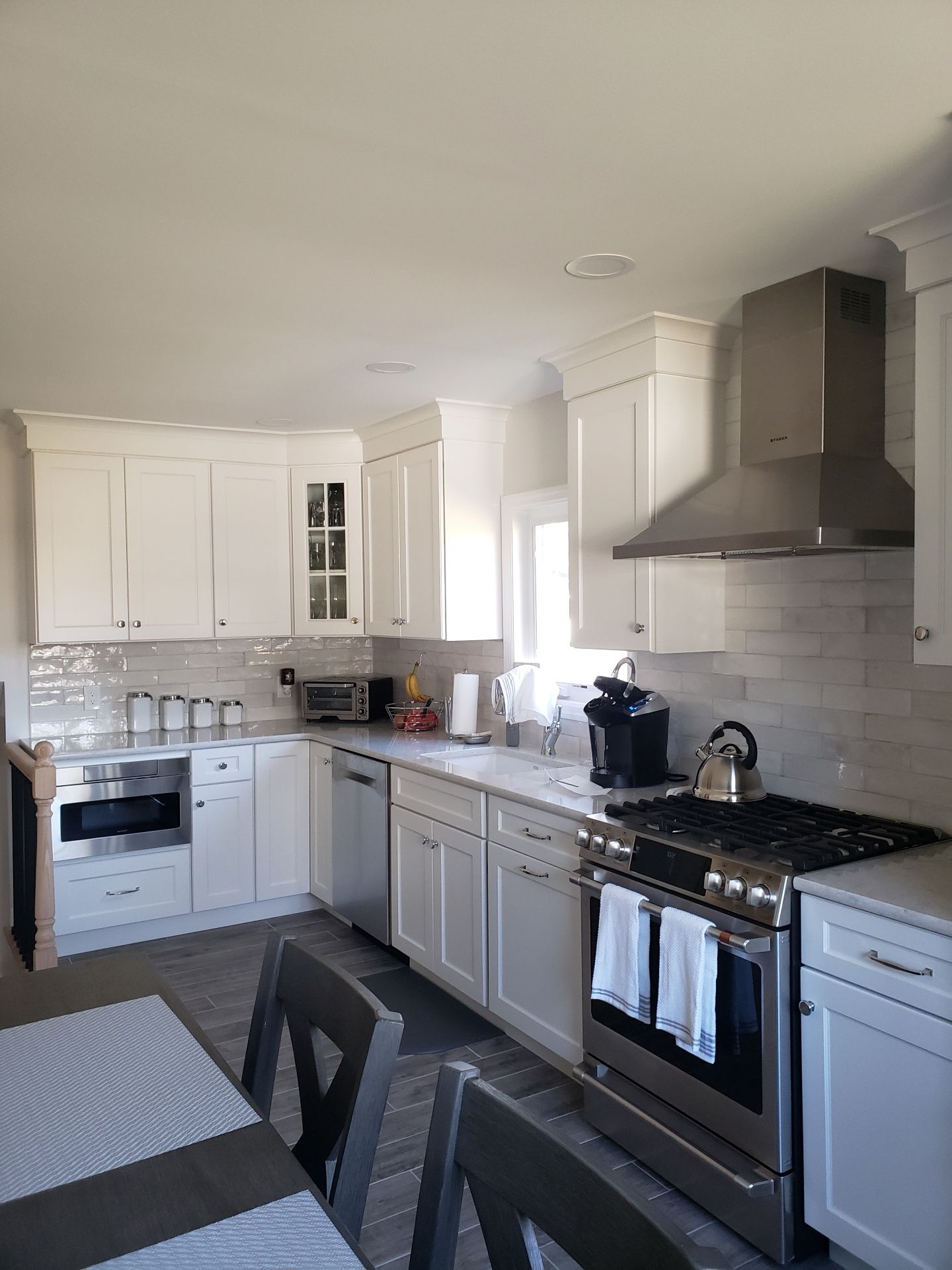 A modern white kitchen featuring white cabinets, stainless steel appliances, a range hood, and a table with chairs.