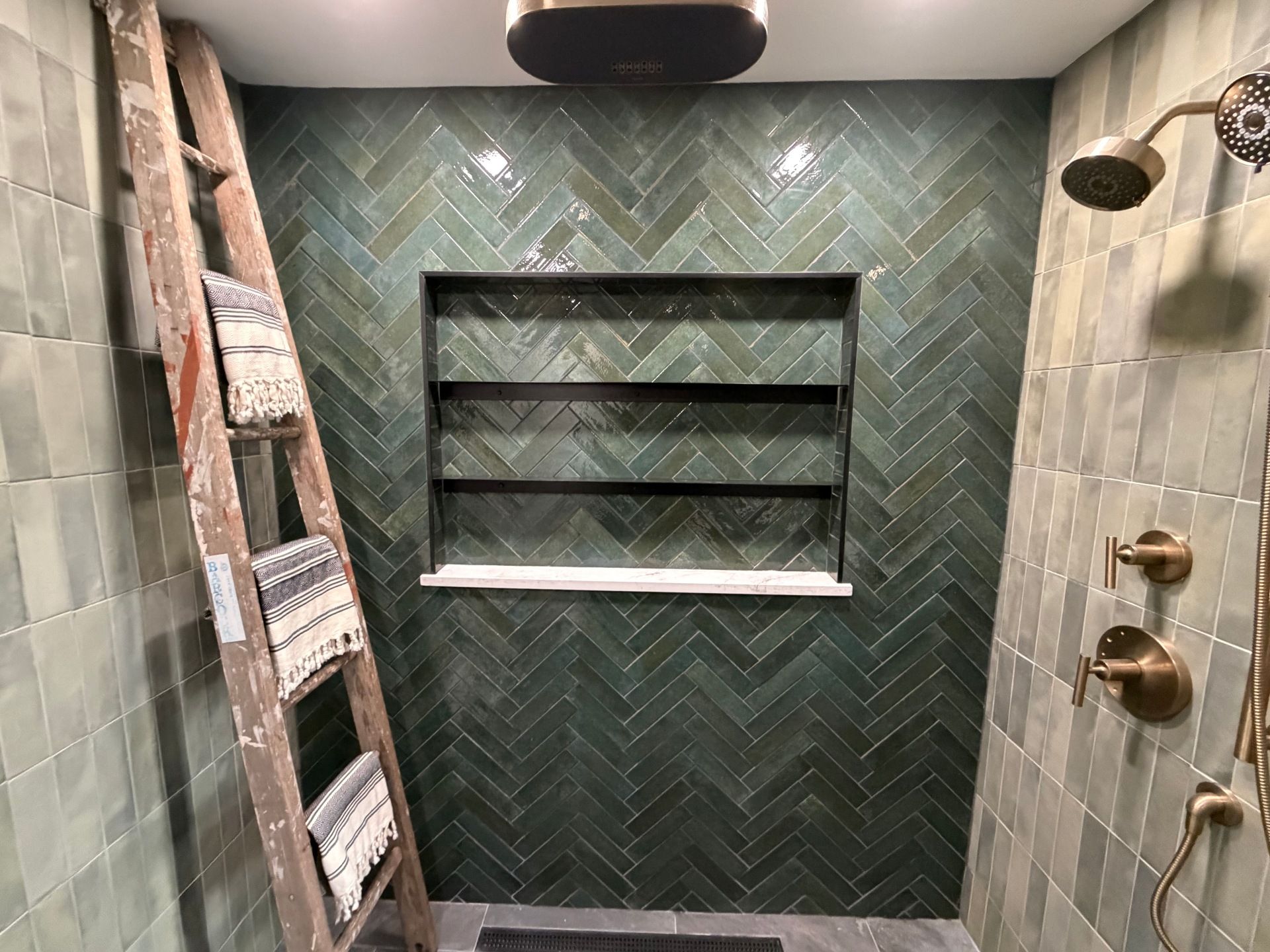A shower with dark green herringbone tile walls, a built-in shelf, brass fixtures, and a decorative wooden ladder.