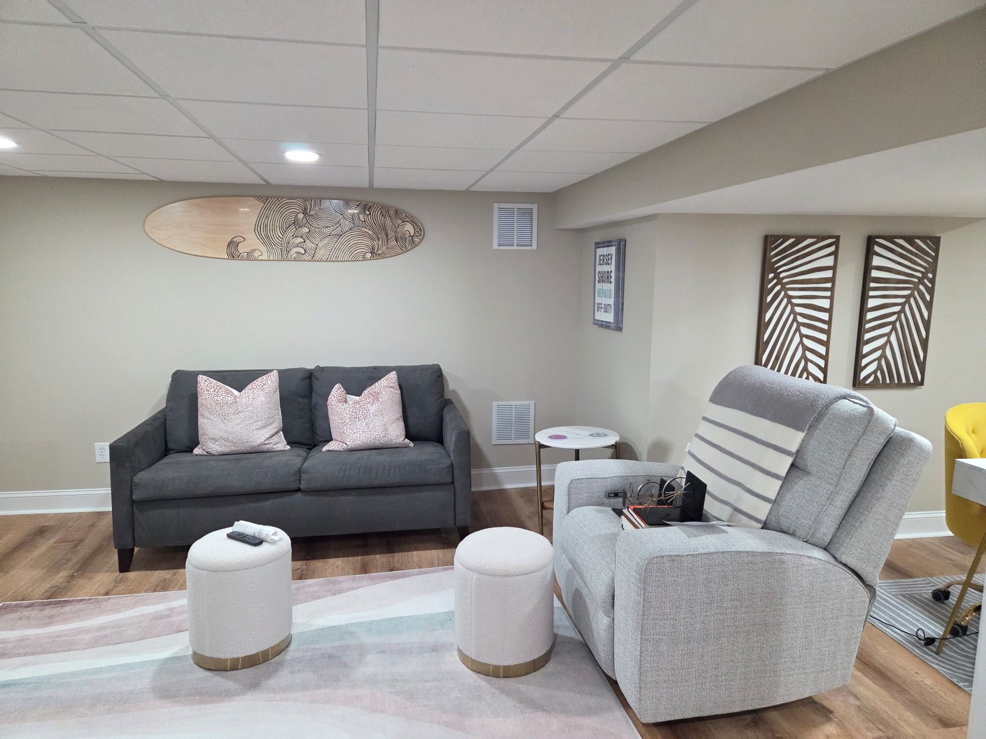 A basement living area with a dark grey sofa, light grey recliner, two white ottomans, and decorative wall art.