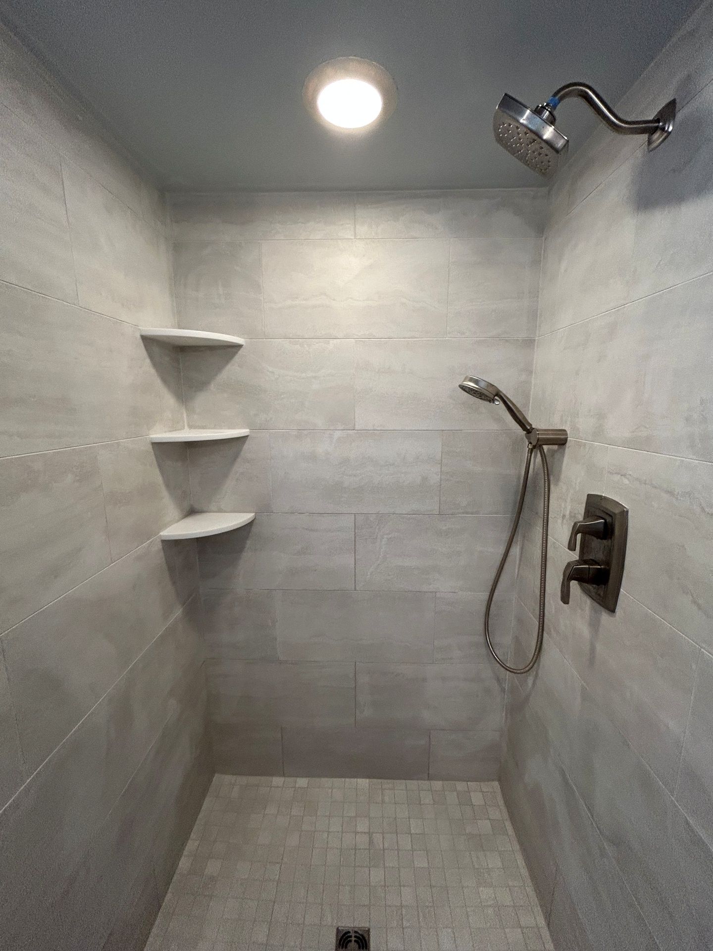 A walk-in shower with light grey tile walls, three corner shelves, a ceiling light, and bronze-finished shower fixtures.