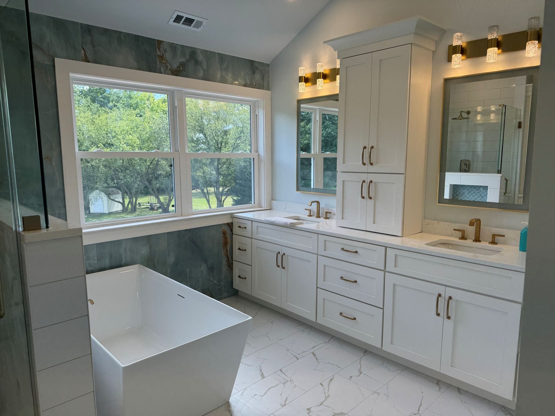 A bright, modern bathroom featuring a white soaking tub, a large window, a white vanity with gold hardware, and mirrors.