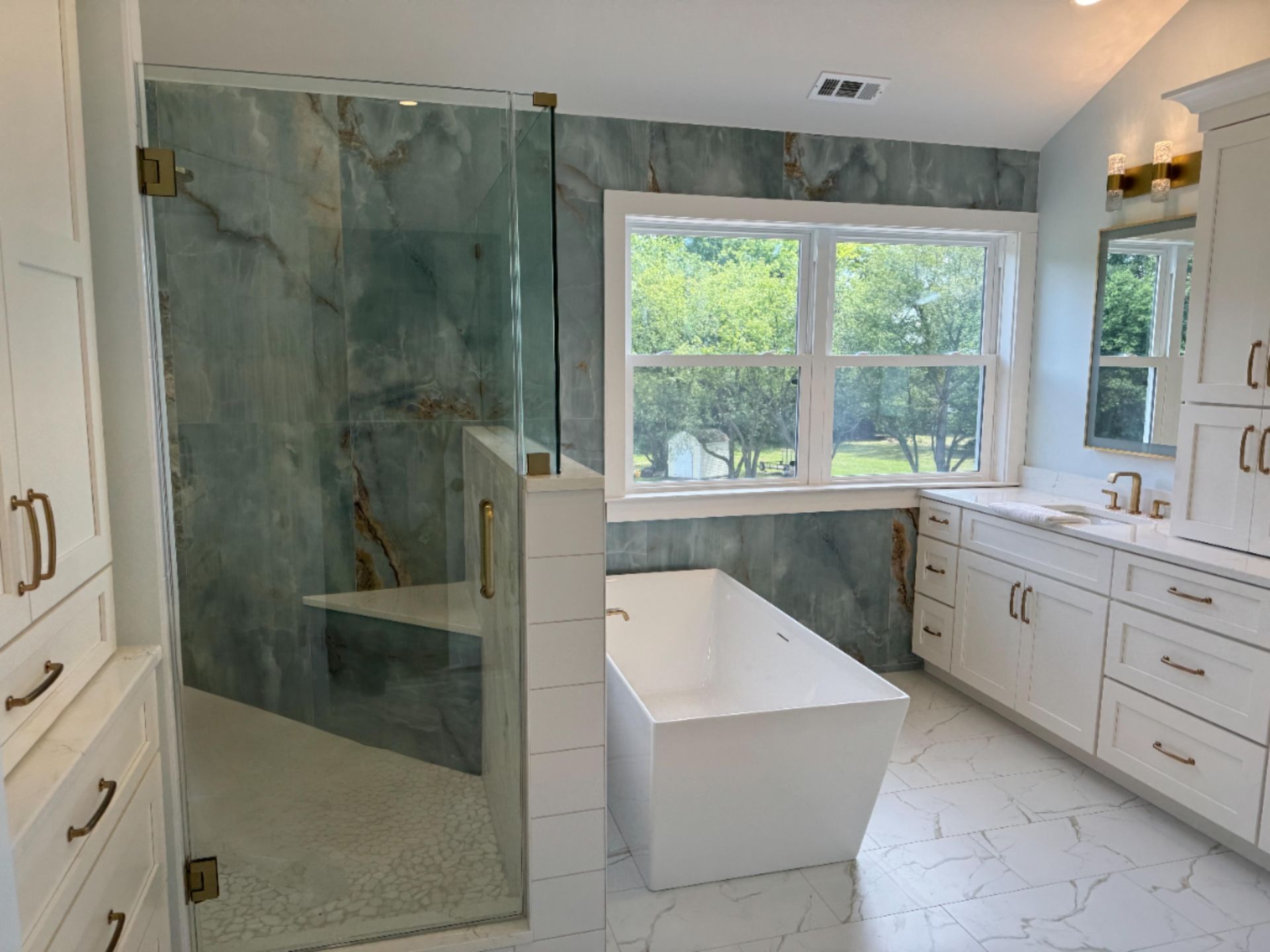 A bright bathroom with a white freestanding tub, a glass-enclosed shower with blue stone tile, and white cabinetry.