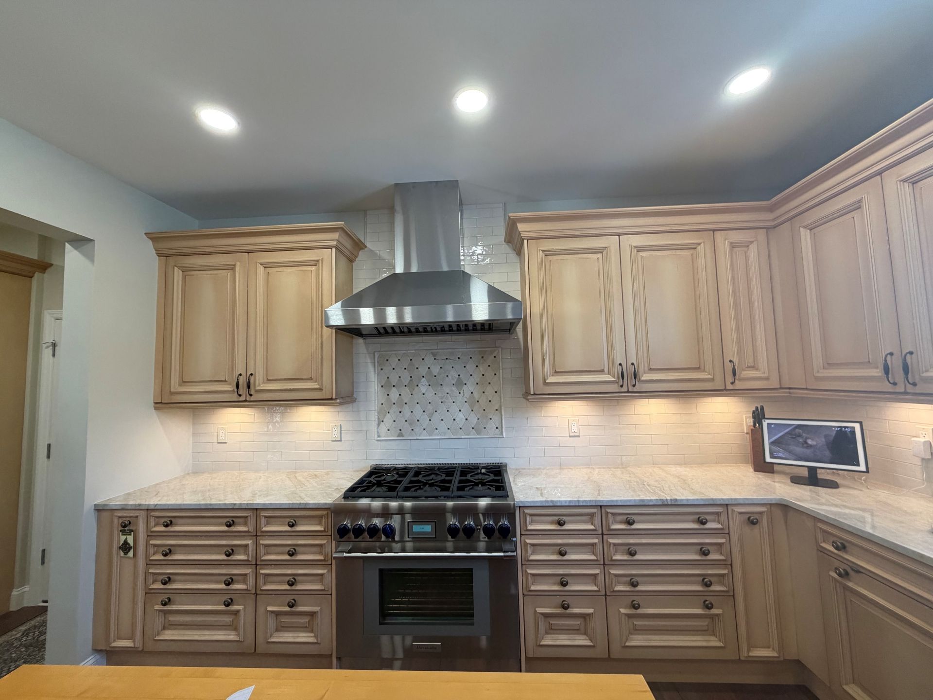 A kitchen with light wood cabinets, stainless steel range and hood, and granite countertops under recessed lighting.