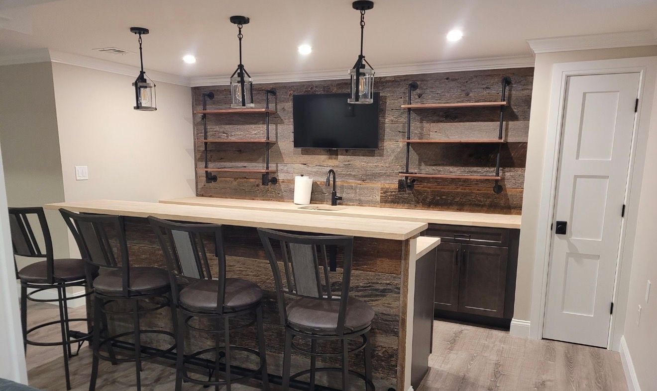 A basement bar with stone walls, wooden shelving, four bar stools, three pendant lights, and a wall-mounted TV.