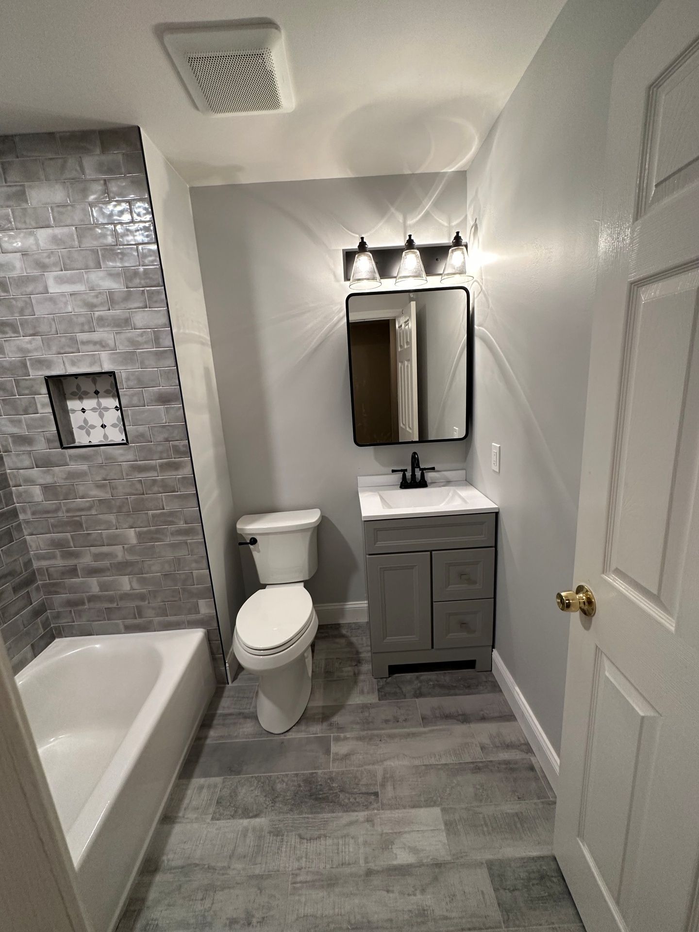 A small bathroom with a gray vanity, mirror, toilet, and a bathtub with a gray-tiled shower wall.