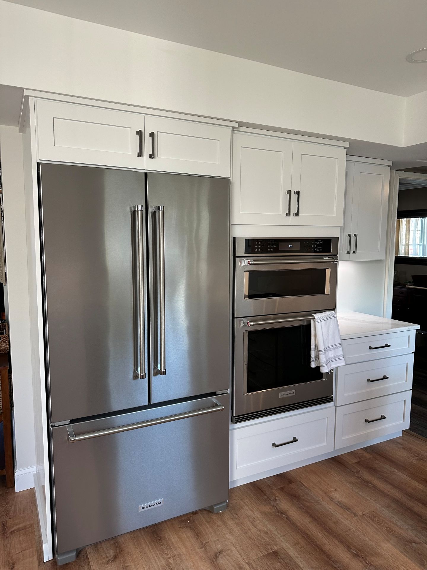 A stainless steel refrigerator next to a built-in double wall oven in a kitchen with white cabinets and wood flooring.