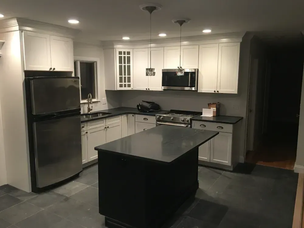 A kitchen with white cabinets, stainless steel appliances, a dark central island, and grey floor tiles.