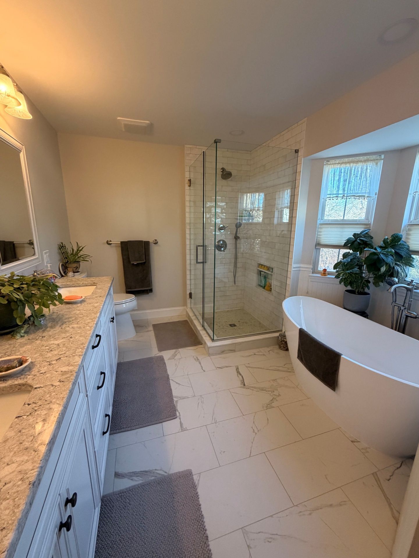A bright bathroom featuring a white vanity with granite countertops, a glass shower, a freestanding tub, and marble floors.