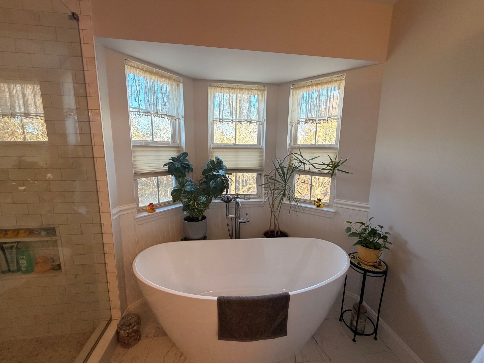 A white freestanding bathtub sits in a bay window nook, flanked by two green potted plants in a modern bathroom.