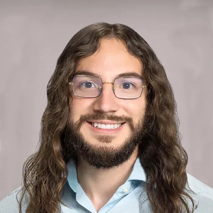 Portrait of a smiling person with long wavy hair, glasses, and a light blue shirt against a gray background