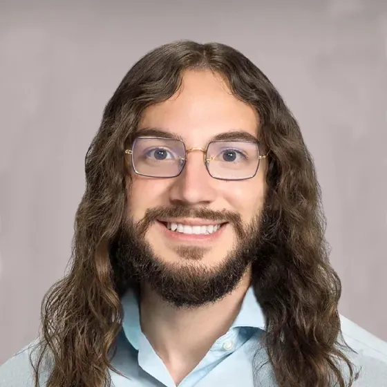 Portrait of a smiling person with long wavy hair, glasses, and a light blue shirt against a gray background