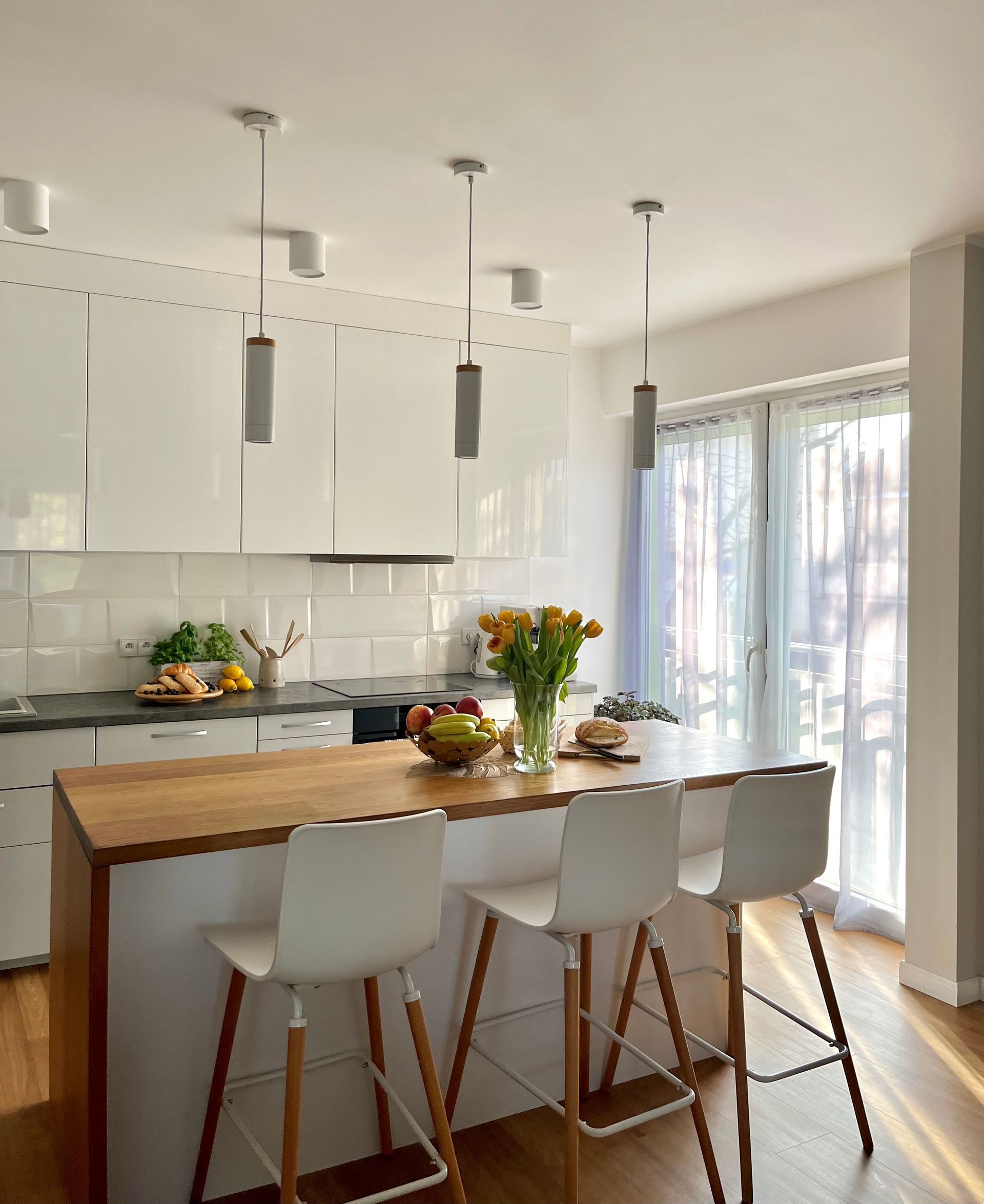 Bright kitchen with a white island, wooden countertop, and bar stools. Cabinets, and a window with sheer curtains.