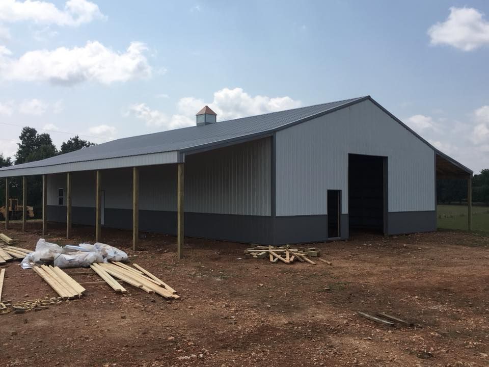 Metal barn under construction with gray and white siding, open doorway, and a covered porch.