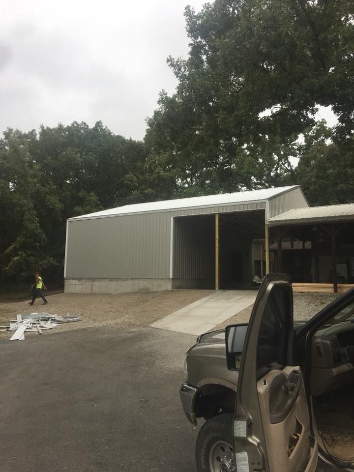 A partially constructed metal building with a concrete ramp and gravel driveway. A person in a high-vis vest walks nearby.