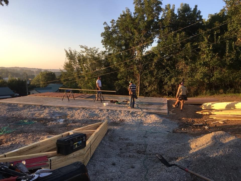 Construction site with three workers on a concrete slab, lumber, tools, and gravel. Green trees and a cityscape in the background.