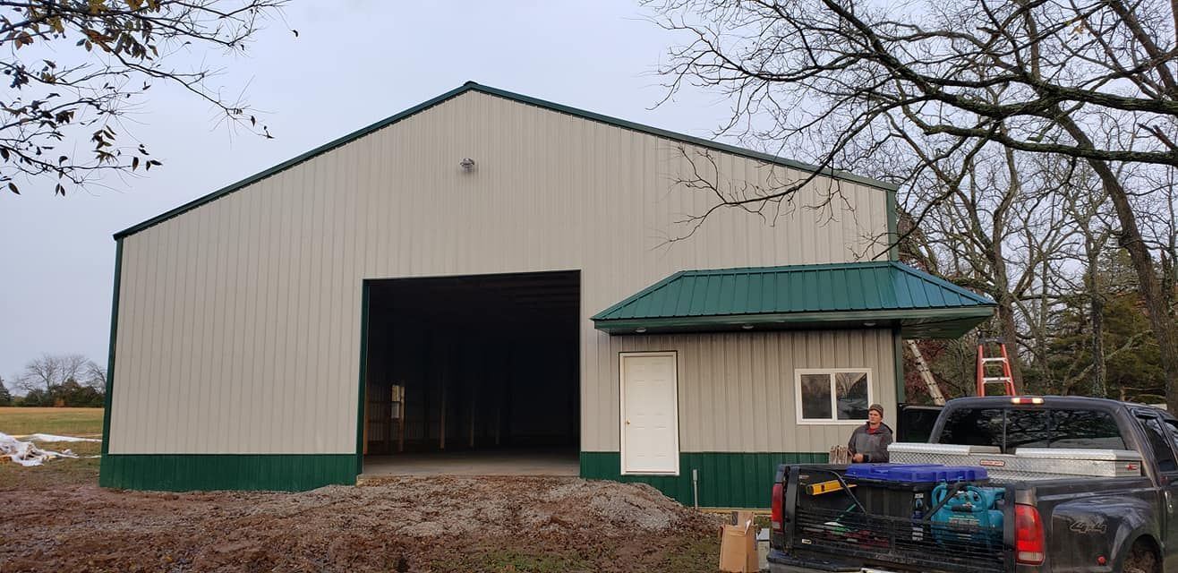 Large tan and green metal building with a truck parked in front. A person is near the truck.