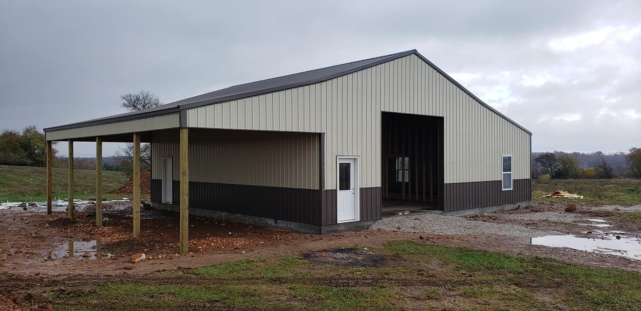 A tan and brown metal barn with a covered area, on a cloudy day, in a field.