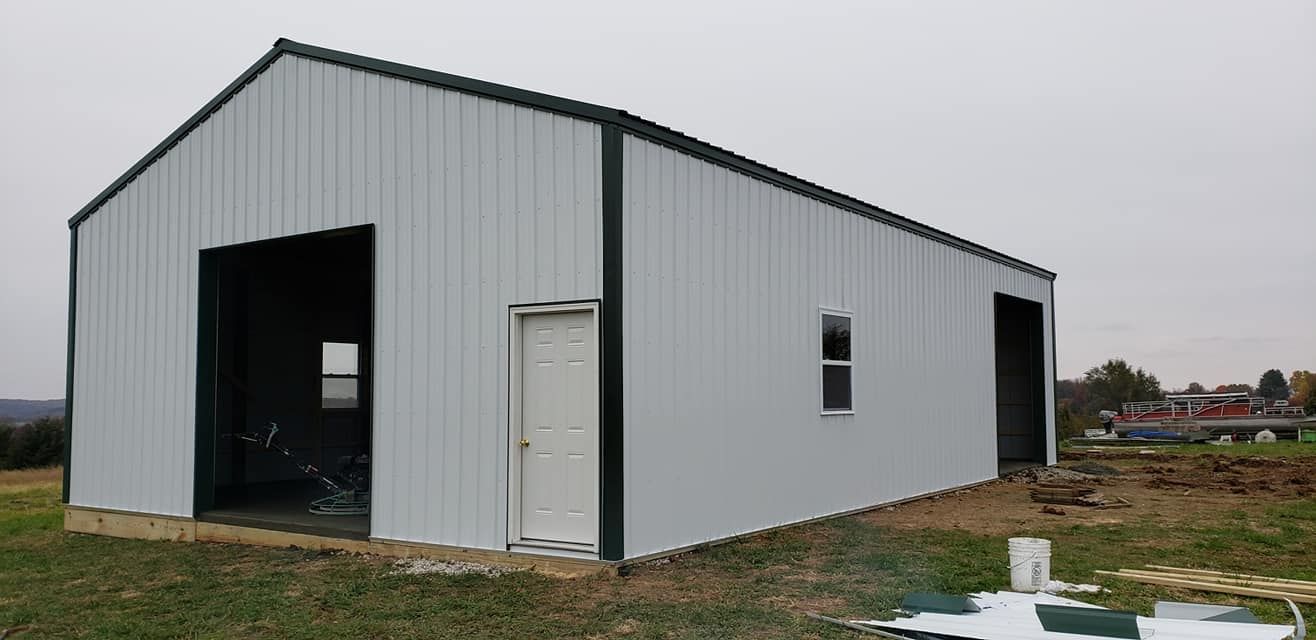 A light gray metal barn with dark green trim and two large openings, set in a grassy field.