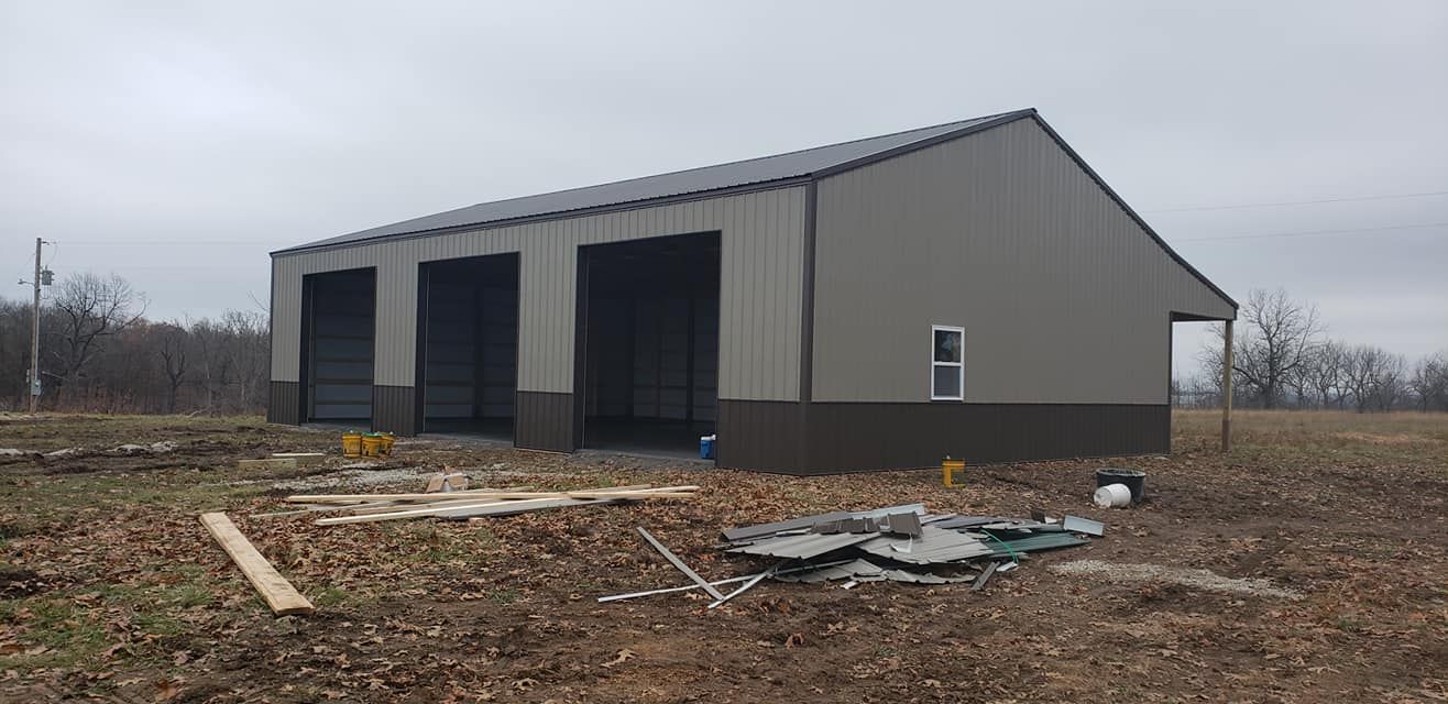 A large, tan metal garage with three bays and a brown base, under construction on a field, overcast sky.
