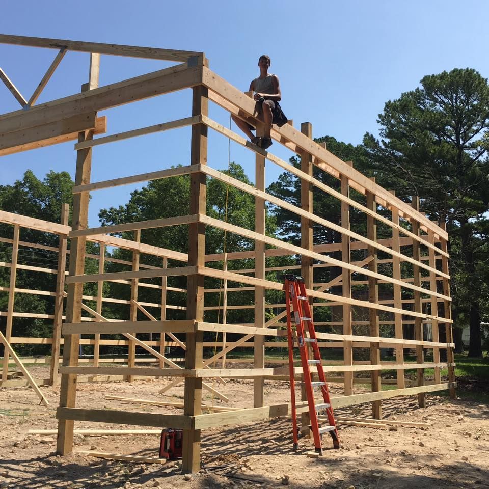 Man on roof of wooden building frame; orange ladder nearby. Bright, sunny day.