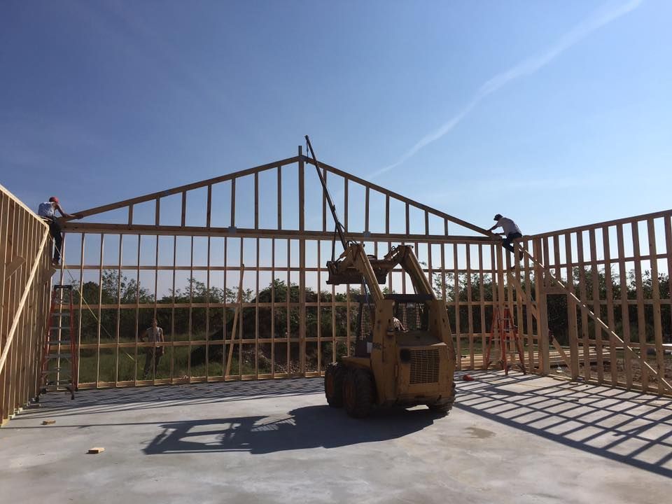 Construction workers placing roof trusses on a new building frame, with a forklift on concrete under a blue sky.