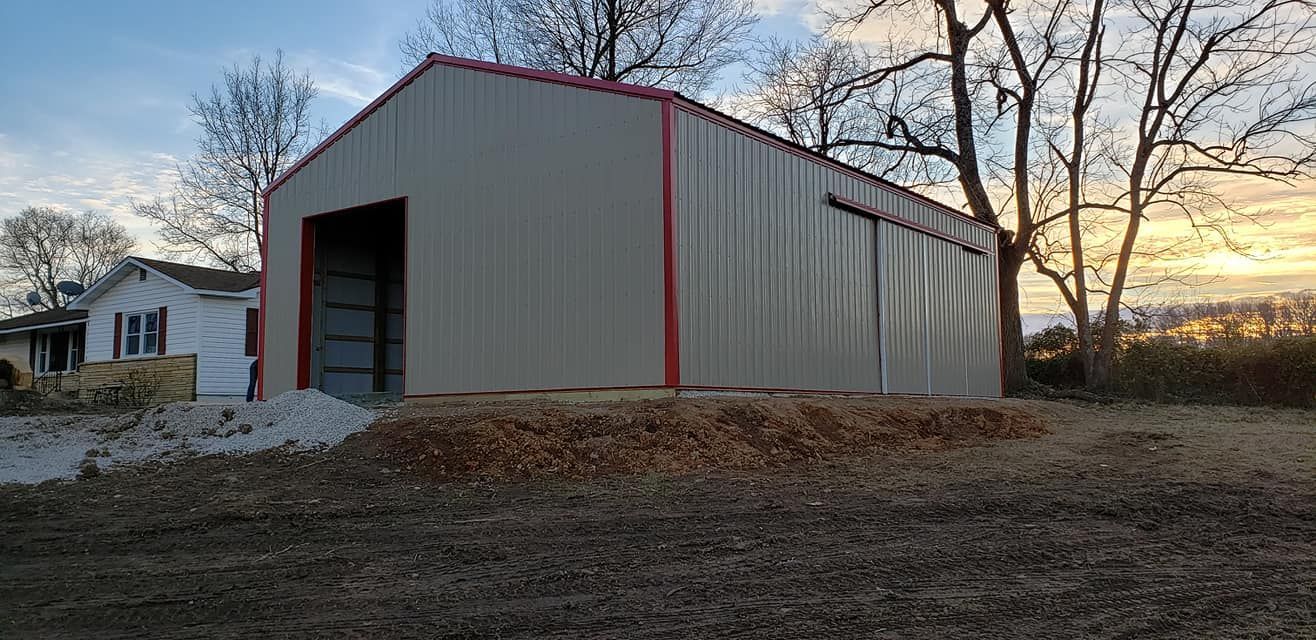 Large metal building with red trim, gravel, and bare trees. A small house is to the left, and a sunset in the background.