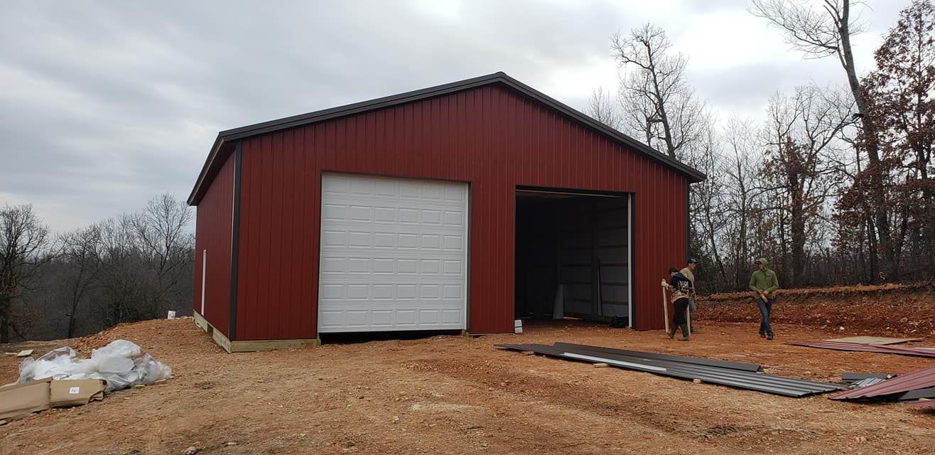Red barn with two garage doors on dirt lot, bare trees in background. Cloudy sky.