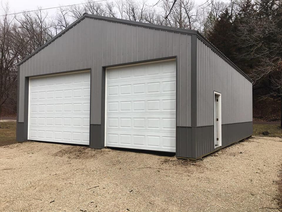 Gray metal garage with two white garage doors and a side door. Gravel driveway.