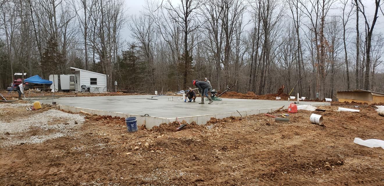 Construction site with a concrete foundation, workers, and a small building in a wooded area.