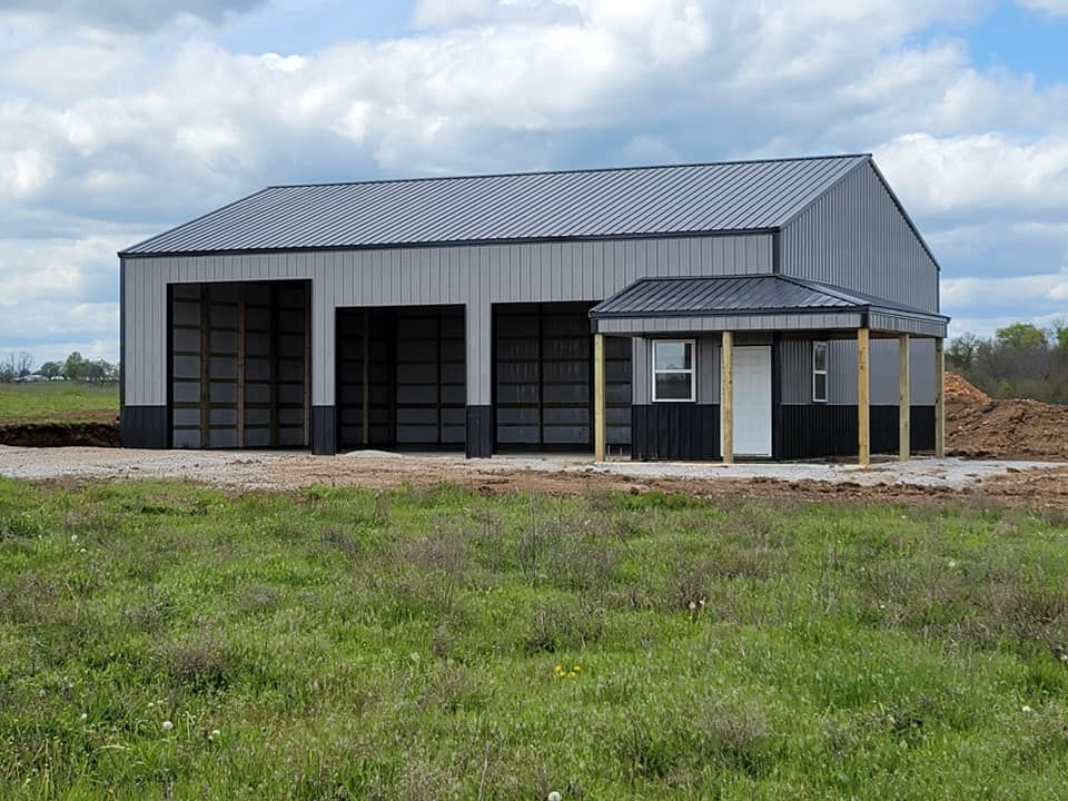 A gray metal building with three garage doors and a small adjoining structure with a window and door, set in a field.