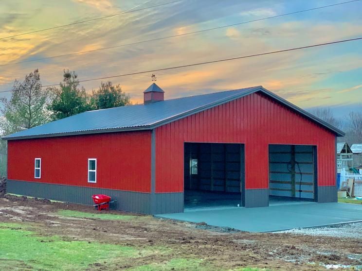 Red metal barn with gray trim, two garage doors, and a concrete pad. Sunset in background.