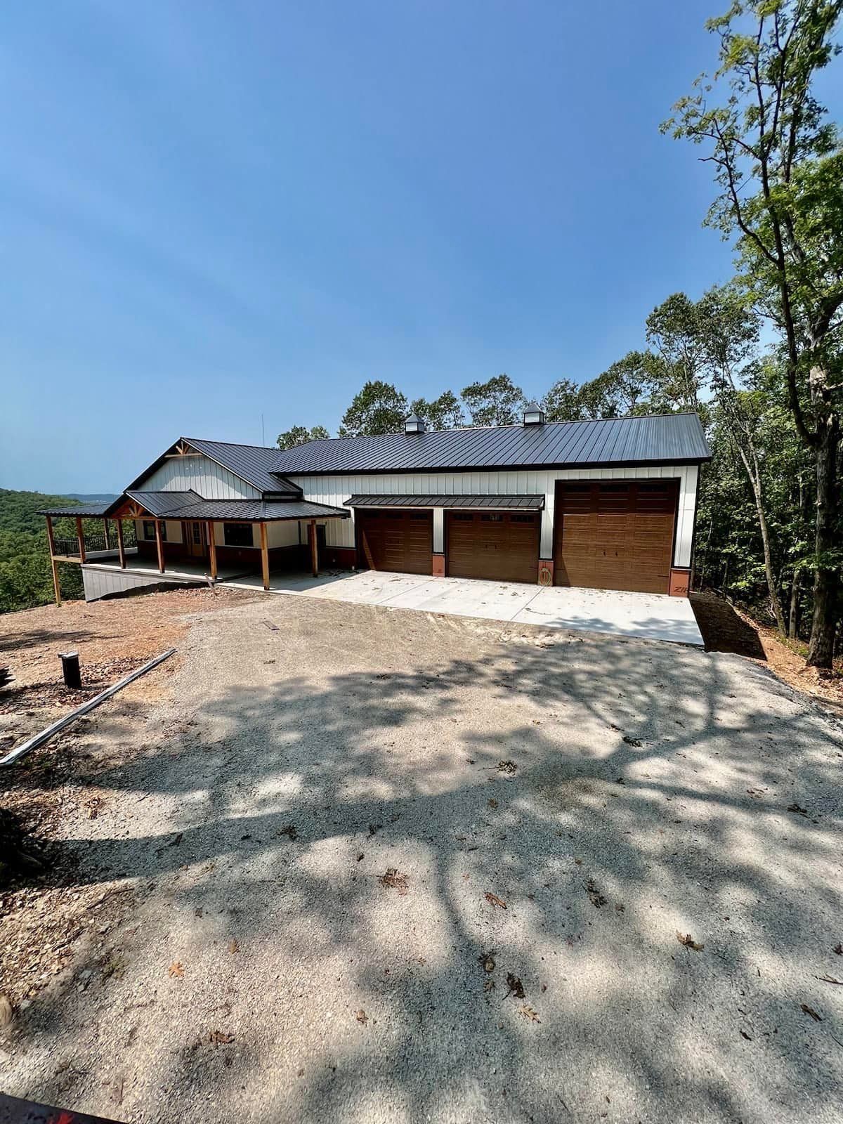 New house with three-car garage and gravel driveway, set against a backdrop of trees and a bright blue sky.