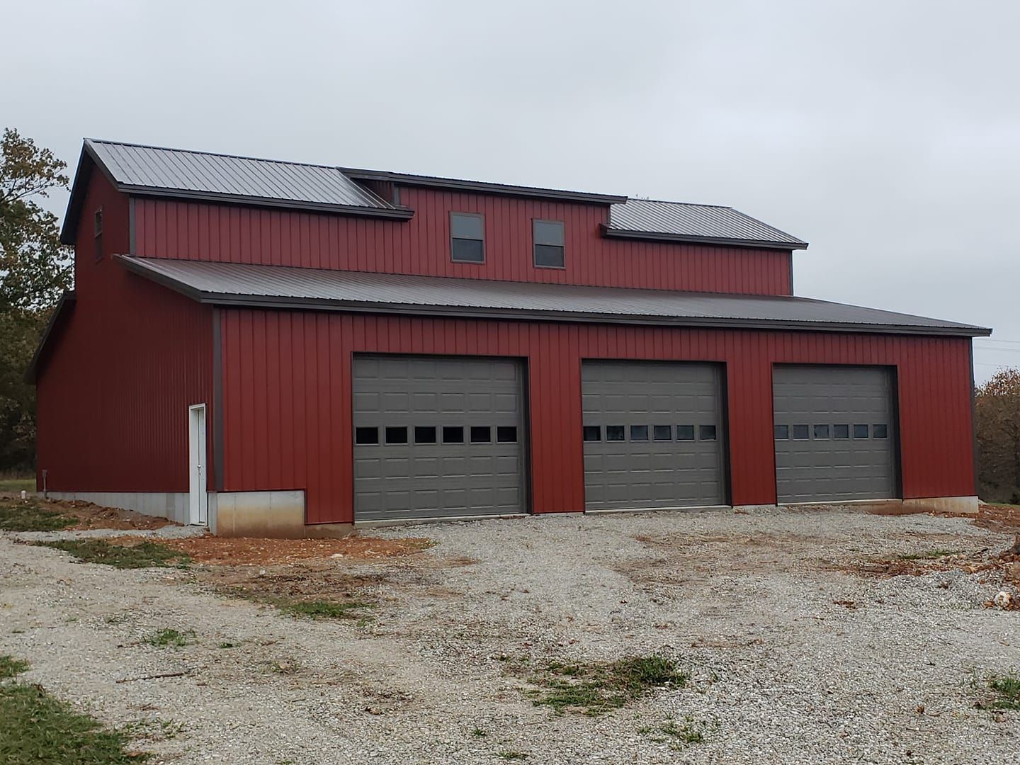 Red metal barn with three garage doors, gray roof, and gravel ground.