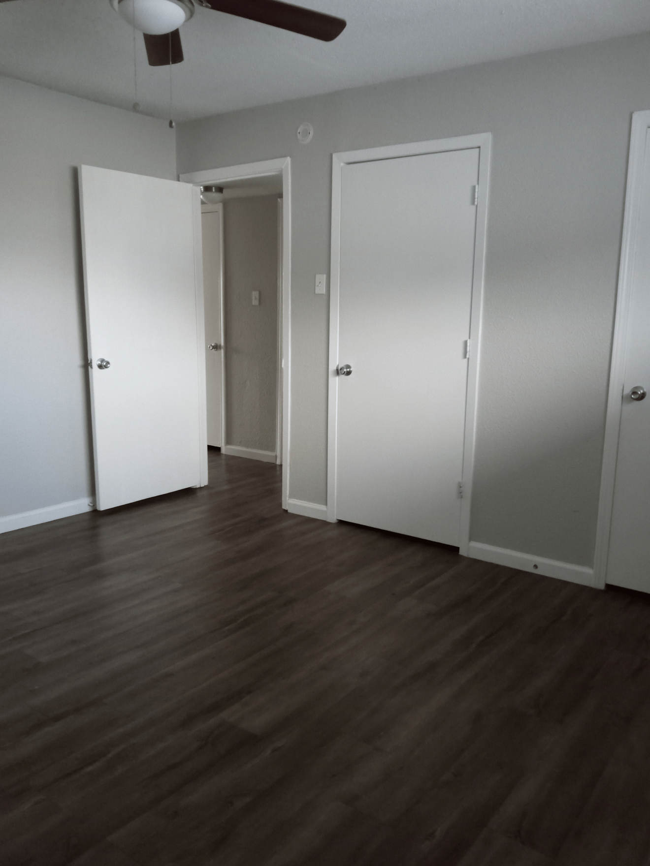 Empty bedroom with three white doors, grey walls, dark wood floor, and a ceiling fan.