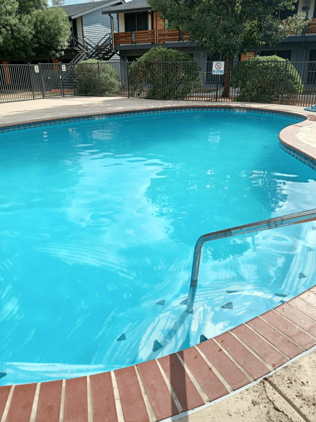 Bright blue swimming pool with brick coping, surrounded by a fence and buildings.