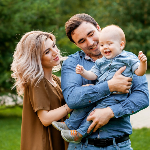 Family of three outdoors: parents holding and smiling at a baby, green background.