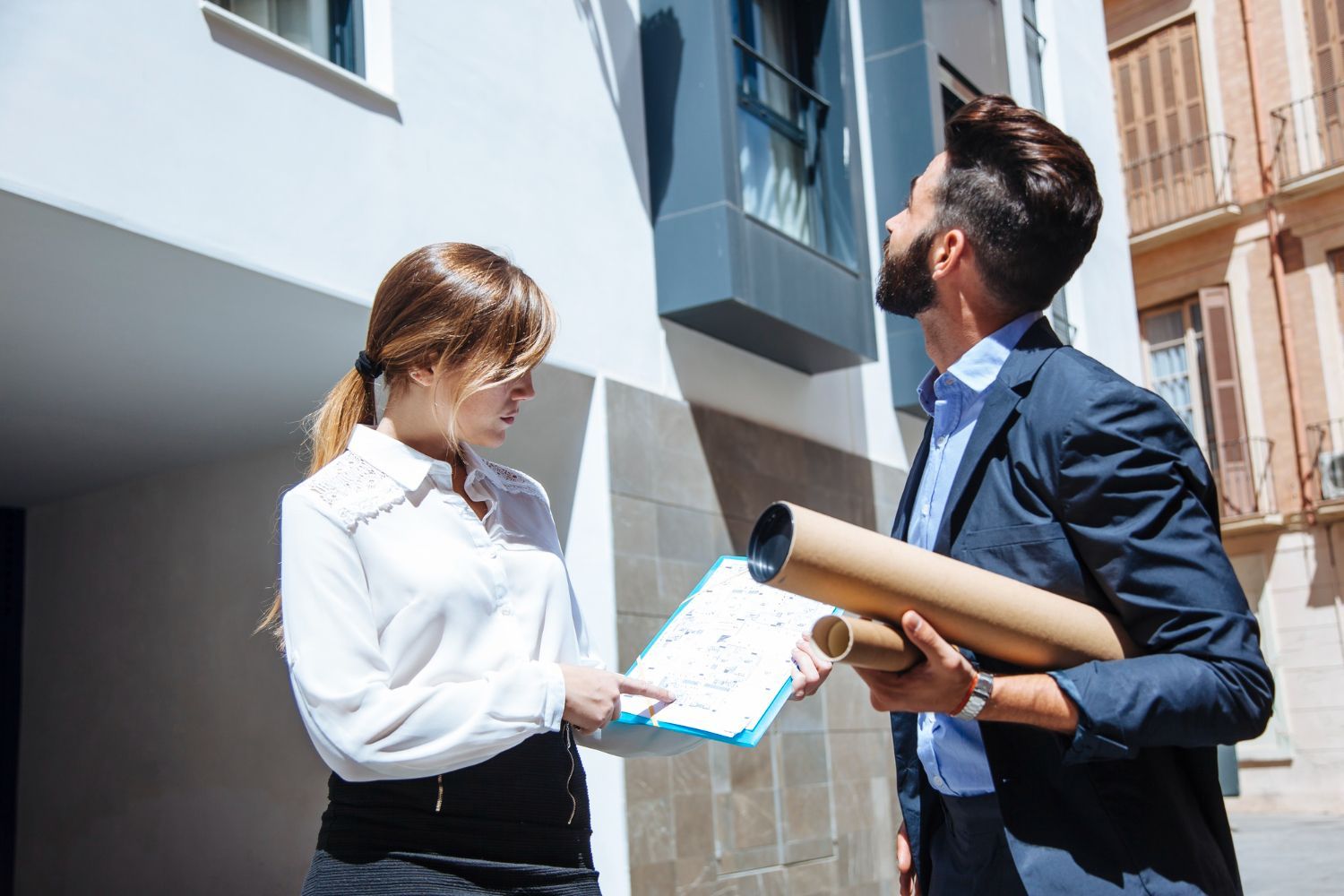 Woman points at a document, man holds blueprints, discussing a building in sunlight.