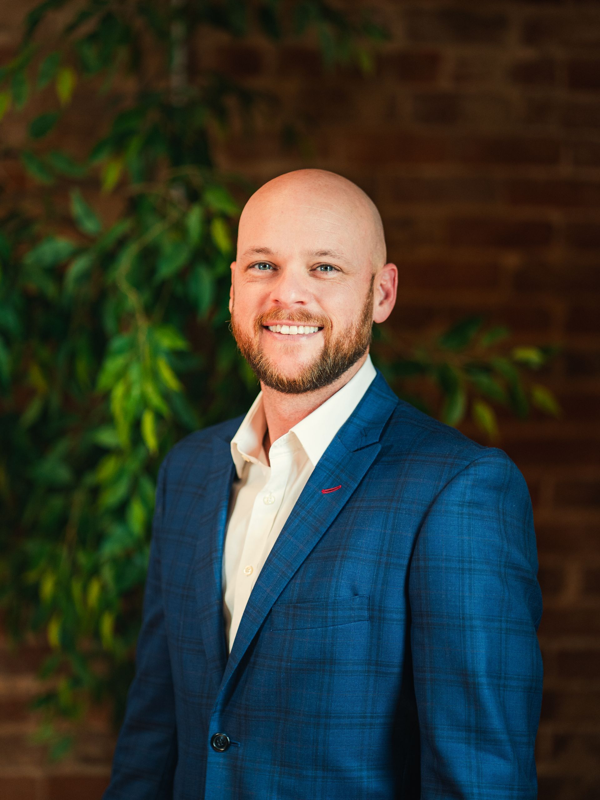 Bald man in glasses smiles, leans against a white wall wearing a blue suit and white shirt.