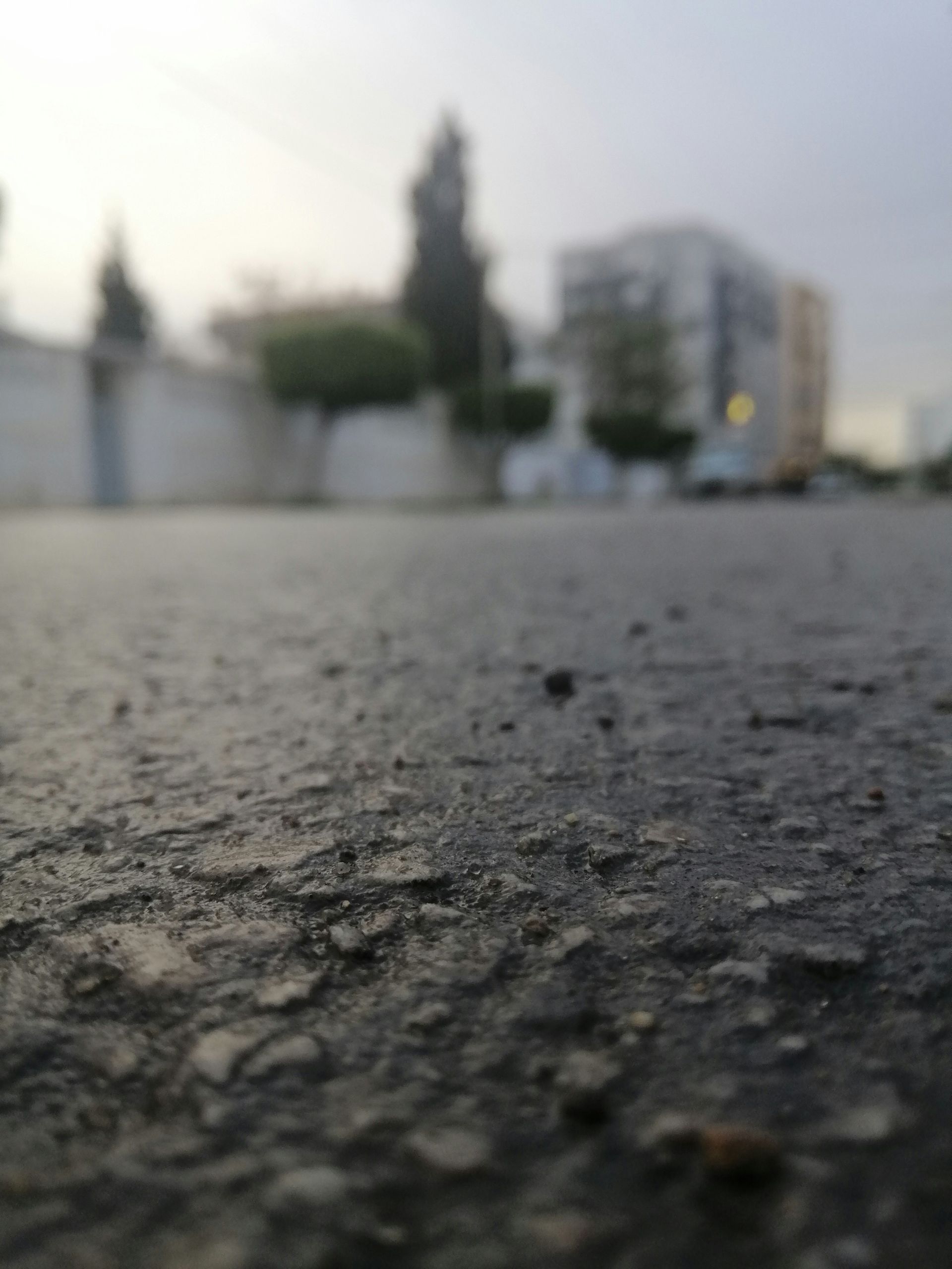 Close-up of asphalt road surface, with blurred buildings and trees in the background under an overcast sky.