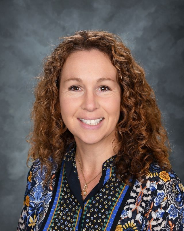 Woman with auburn curly hair, smiling, wearing a dark burgundy shirt, against a mottled gray background.