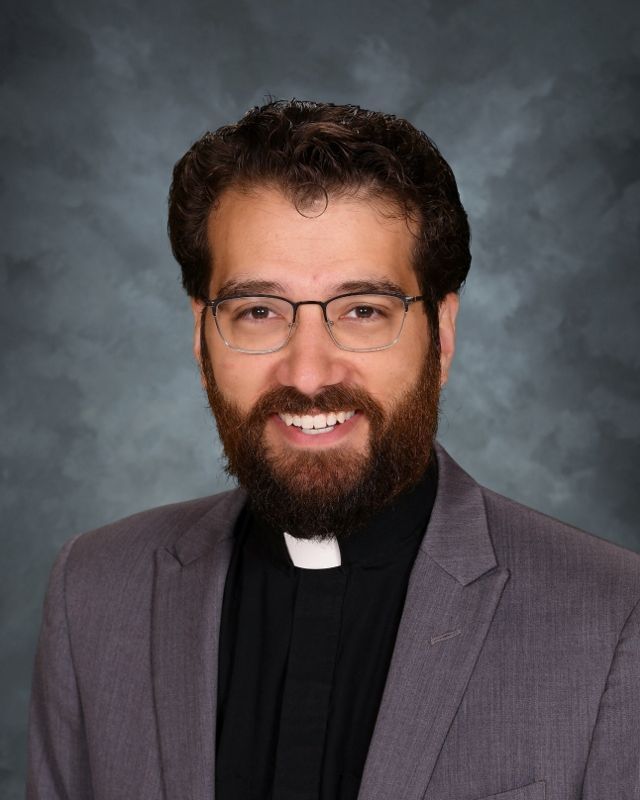 Clerical man with glasses and beard, wearing a black suit and collar, smiling.