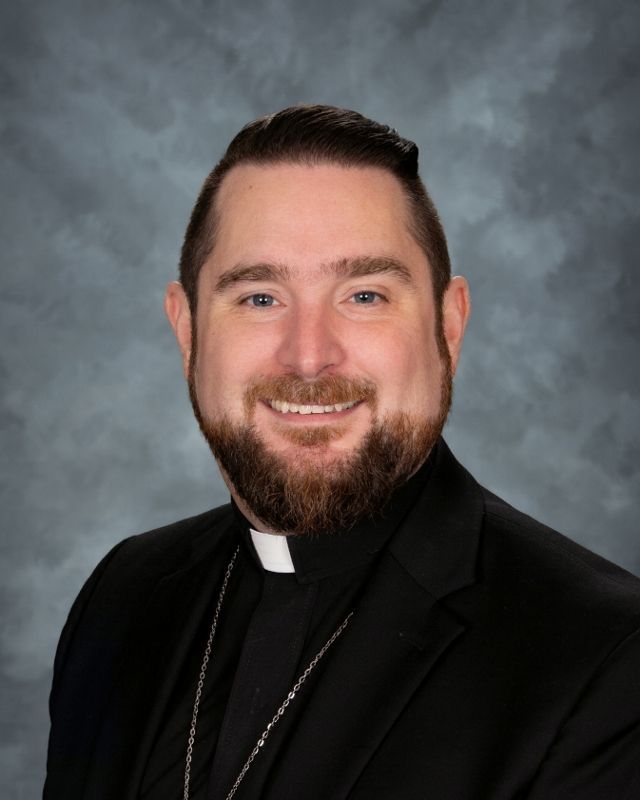 Man with glasses and beard, wearing clerical collar and gray blazer, smiles.